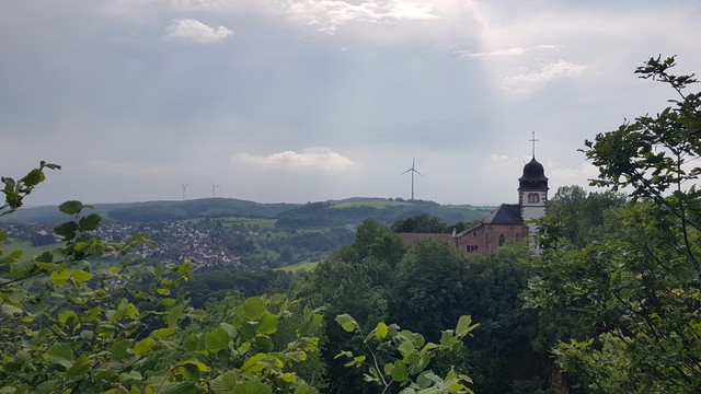 Grüne Landschaft im Mittelgebirge. Rechts im Bild steht eine Kirche auf einem Berg. Links liegt ein Dorf im Tal.