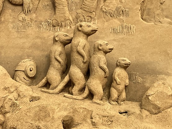 Eine aus Sand gestaltete Erdmännchenfamilie, dahinter steht ein Minion
