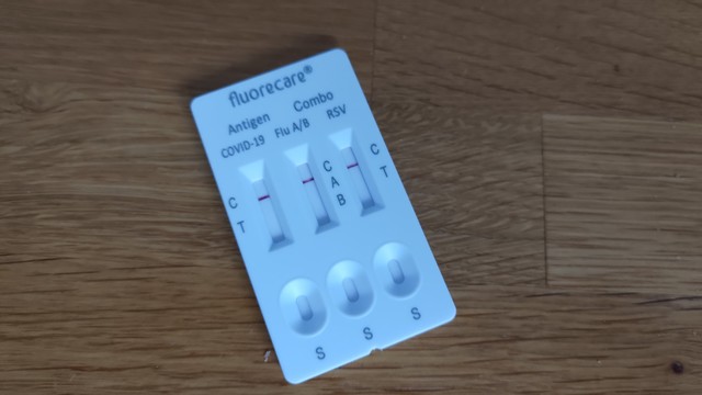 Foto eines lateral flow tests, auch Schnelltest genannt. Ein weißes Plastik-Rechteck mit drei winzigen Trichtern und drei Papierstreifen. Beschriftet mit dem Herstellernamen fluorecare und Antigen Combo COVID-19, Flu A/B, RSV.

Der Test ist bereits abgeschlossen und zeigt bei jedem der Tests den 